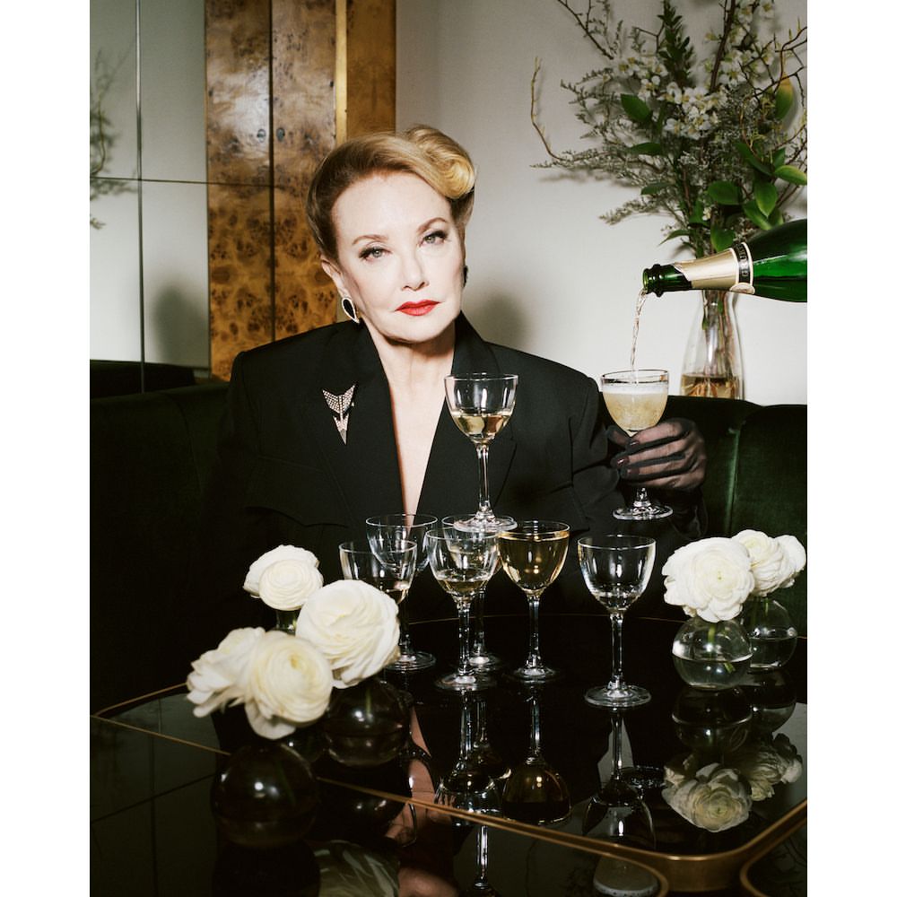 Elegant mature woman in a black blazer poses at a table with champagne glasses as champagne is poured, accented by flowers and a stylish backdrop.