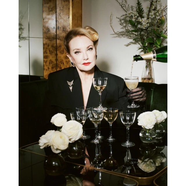 Elegant mature woman in a black blazer poses at a table with champagne glasses as champagne is poured, accented by flowers and a stylish backdrop.