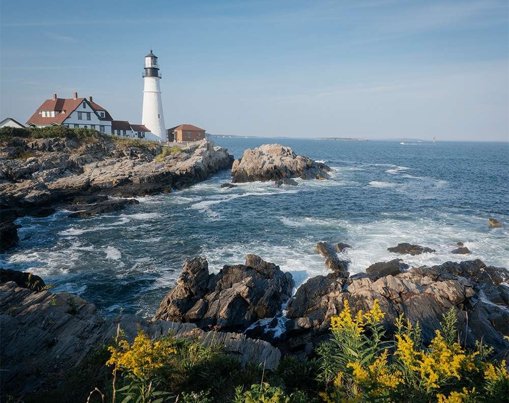Scenic view of the Portland Head Light lighthouse on a rocky coastline with the Atlantic Ocean under a clear blue sky.