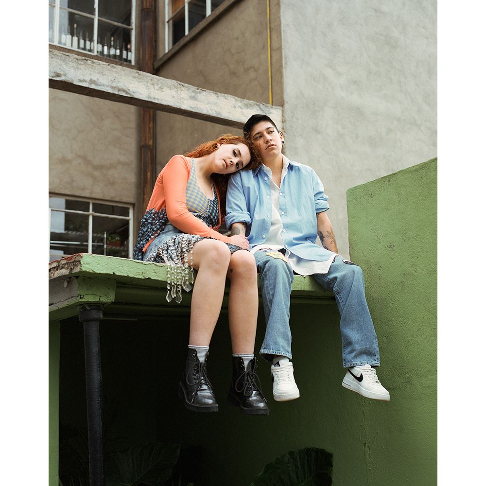 Two young adults, one with red hair and the other with short dark hair, sit side-by-side on a rooftop ledge against a concrete background.