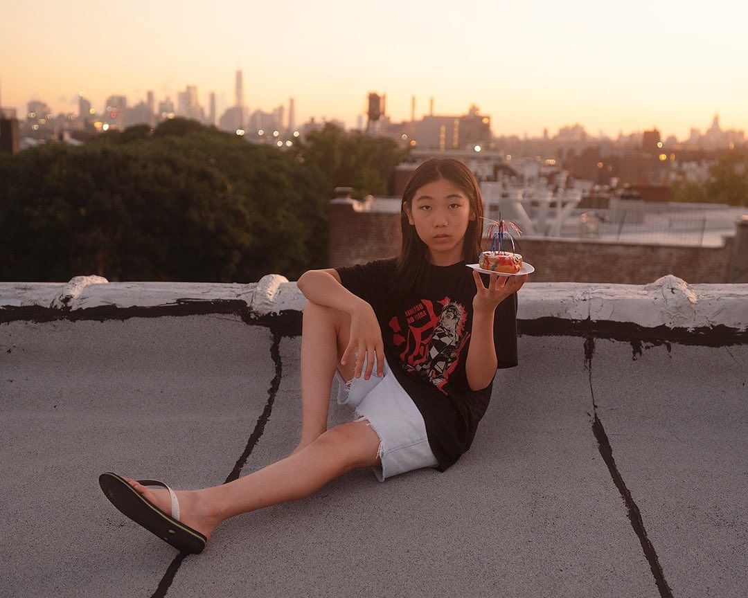 Asian female sitting on a rooftop holding a cake at sunset with a cityscape in the background, wearing a graphic tee and jean shorts.