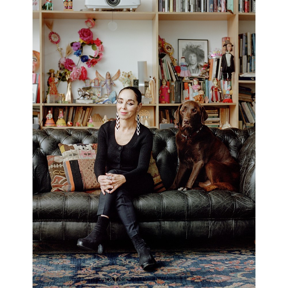 Adult woman sits on a leather couch with a brown Labrador dog indoors with a bookshelf, books, and art in the background.