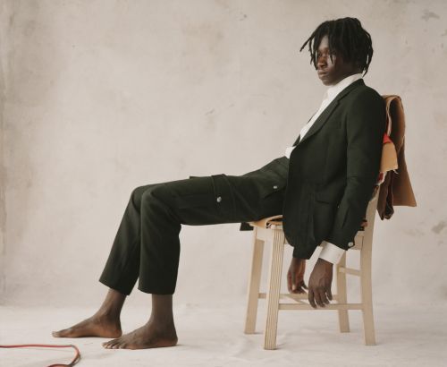 Barefoot male model in dark green suit sits on a wooden chair in a neutral-toned studio, striking a relaxed pose with a contemplative expression.