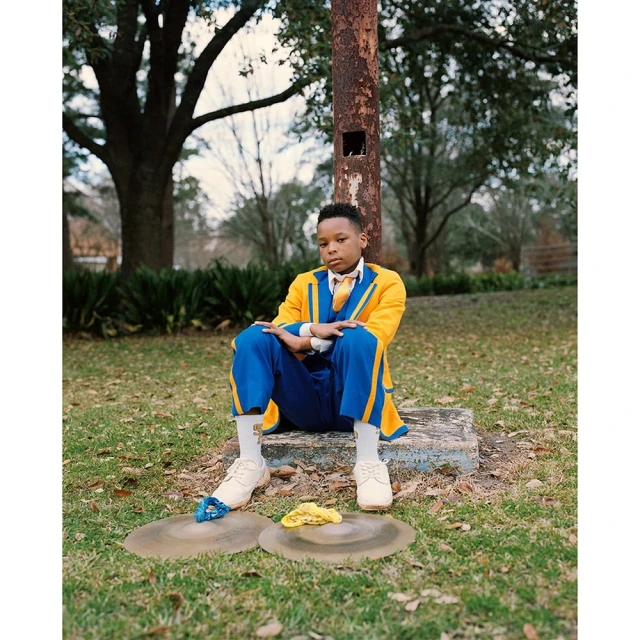 Boy in blue and yellow suit sits on concrete near cymbals in outdoor setting, with trees in the background, captured in medium shot.