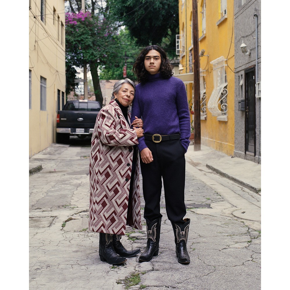 Woman and young adult stand together in an alleyway, both wearing boots, in a medium shot with yellow buildings as background.