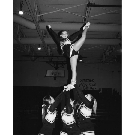 Group of cheerleaders performing a stunt indoors. One cheerleader is elevated and balanced by her teammates in black and white.