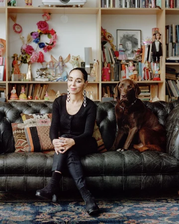Adult woman with a dog sits on a black leather couch in a room with a bookshelf filled with books and collectibles.