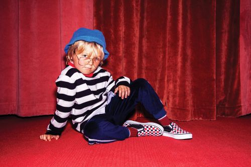 Young child with blond hair and glasses poses on a red floor in front of red curtains, wearing striped shirt, blue hat and checkerboard shoes.