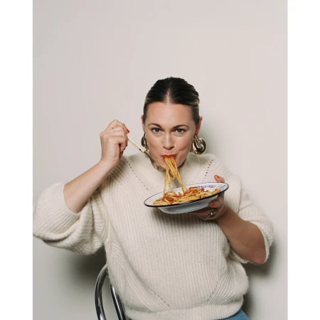 Woman in cream sweater holding a bowl and eating spaghetti with a fork against a neutral background