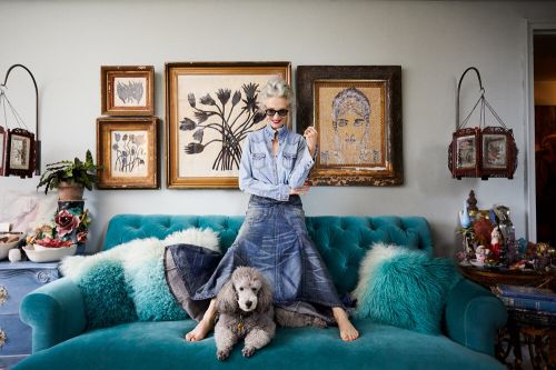 An elderly woman with grey hair wearing a denim shirt and skirt seated on a teal tufted sofa with a poodle.