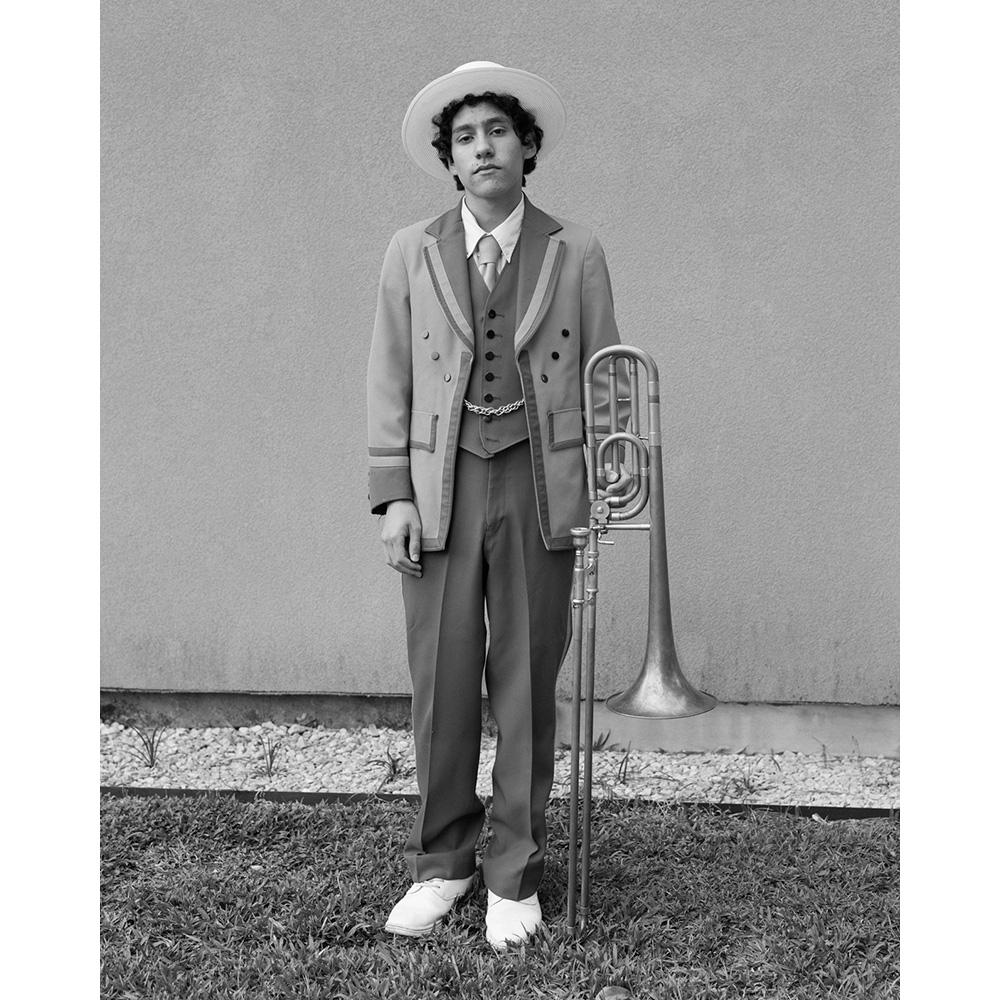 Monochrome portrait of a young man in vintage suit with a fedora, standing outdoors with a trombone against a neutral wall.