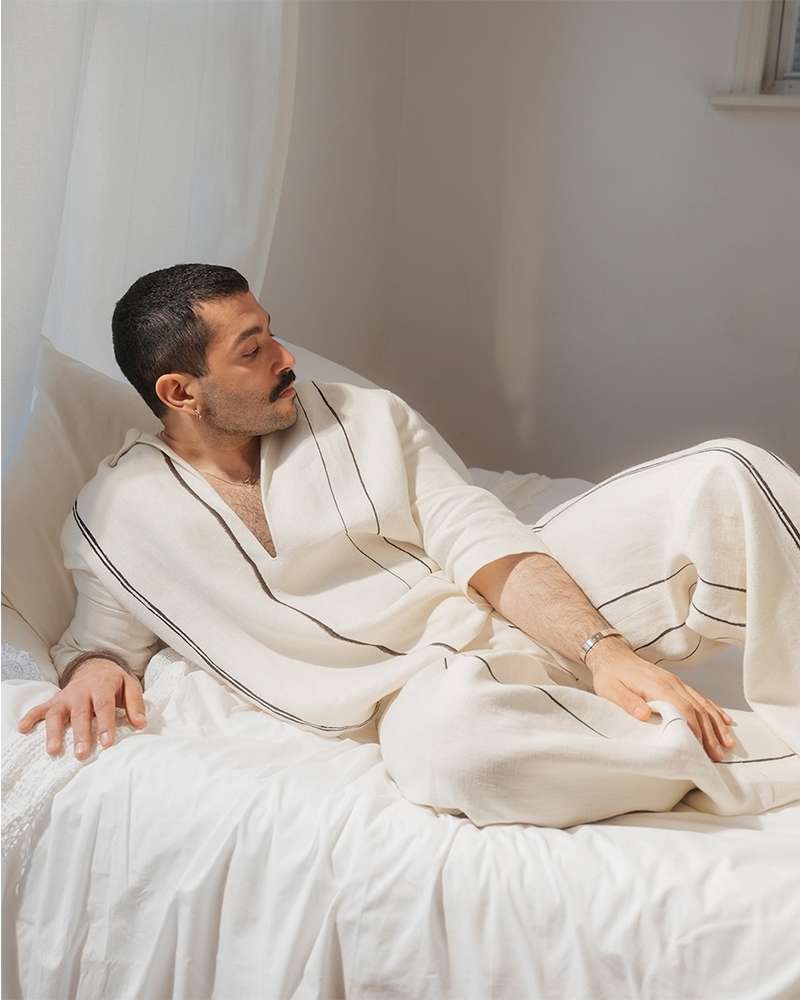Man in relaxed pose on white bed wearing white linen pyjamas with black trim, looking off to the side in natural light.
