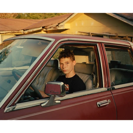 Young man in a black t-shirt seated inside a vintage maroon car parked outdoors in front of a house