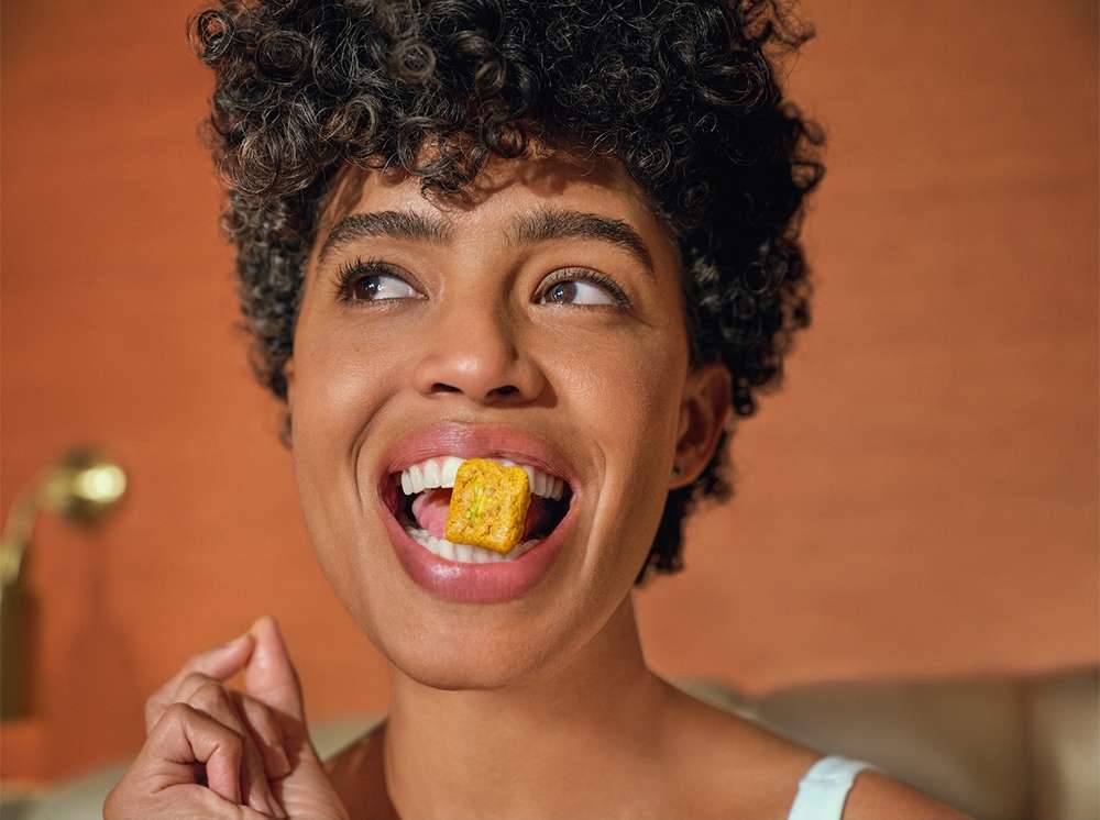 Close-up portrait of a smiling woman with curly hair, holding a square, yellow food item in her mouth. Studio lighting. 