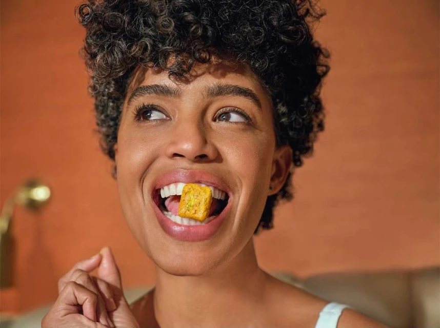 Close-up portrait of a smiling woman with curly hair, holding a square, yellow food item in her mouth. Studio lighting. 