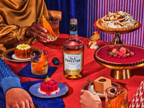 Festive table arrangement with bottle of Old Forester bourbon, desserts, and cocktails, set against red striped backdrop.