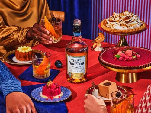 Festive table arrangement with bottle of Old Forester bourbon, desserts, and cocktails, set against red striped backdrop.