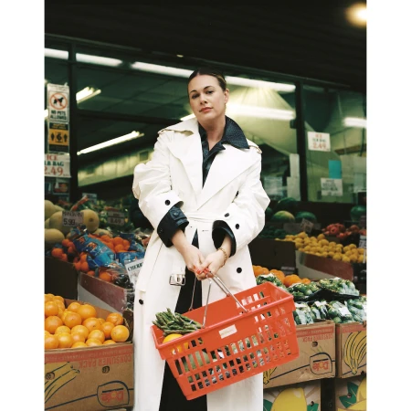 Adult woman in white trench coat holds orange shopping basket outside a fruit and vegetable market with fresh produce.