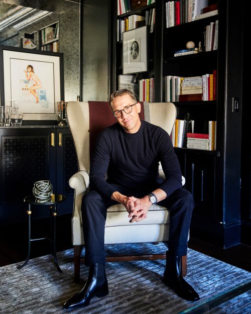 Stylish man sits in a wingback chair in a room with a bookcase, wearing a black sweater, black pants, and black boots.