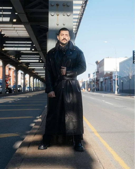 Man in long black leather coat stands beneath an elevated structure on a city street, eyes closed, bathed in natural light.