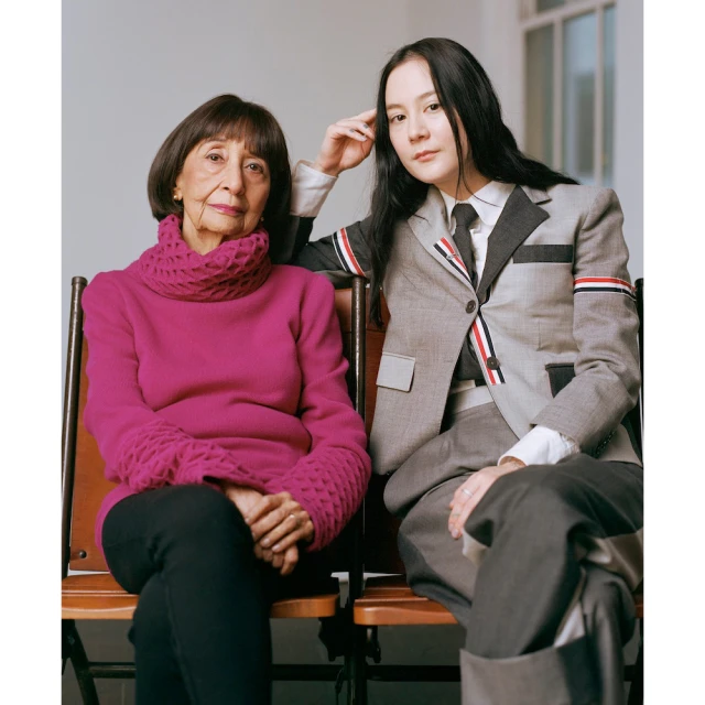 Elegant portrait of two women seated together, one in a pink sweater and the other in a grey suit, set against a neutral backdrop.
