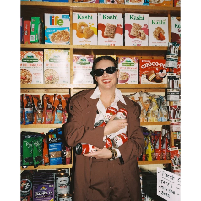 A woman in a brown suit smiles while holding Campbell's soup cans, standing in front of a grocery shelf filled with cereal and snacks.