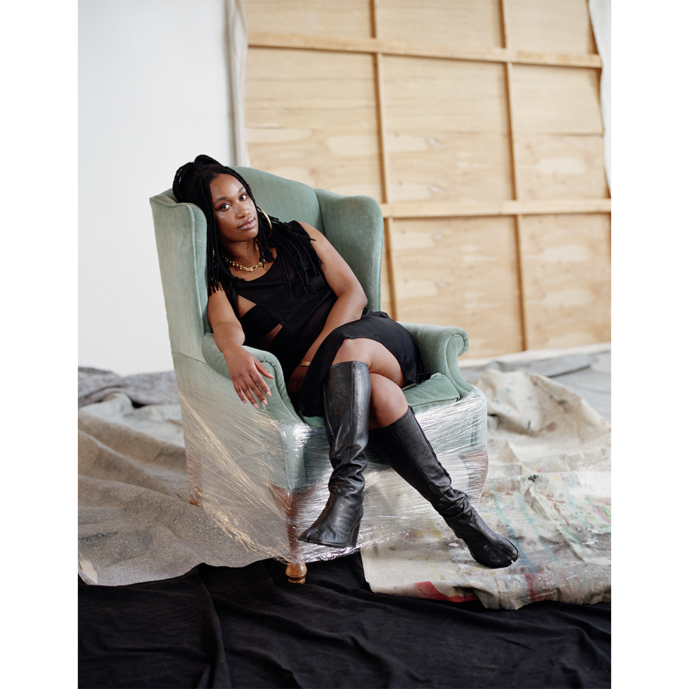 An African American woman in a black dress and boots is seated in a vintage chair wrapped in plastic in an indoor photo studio setting.