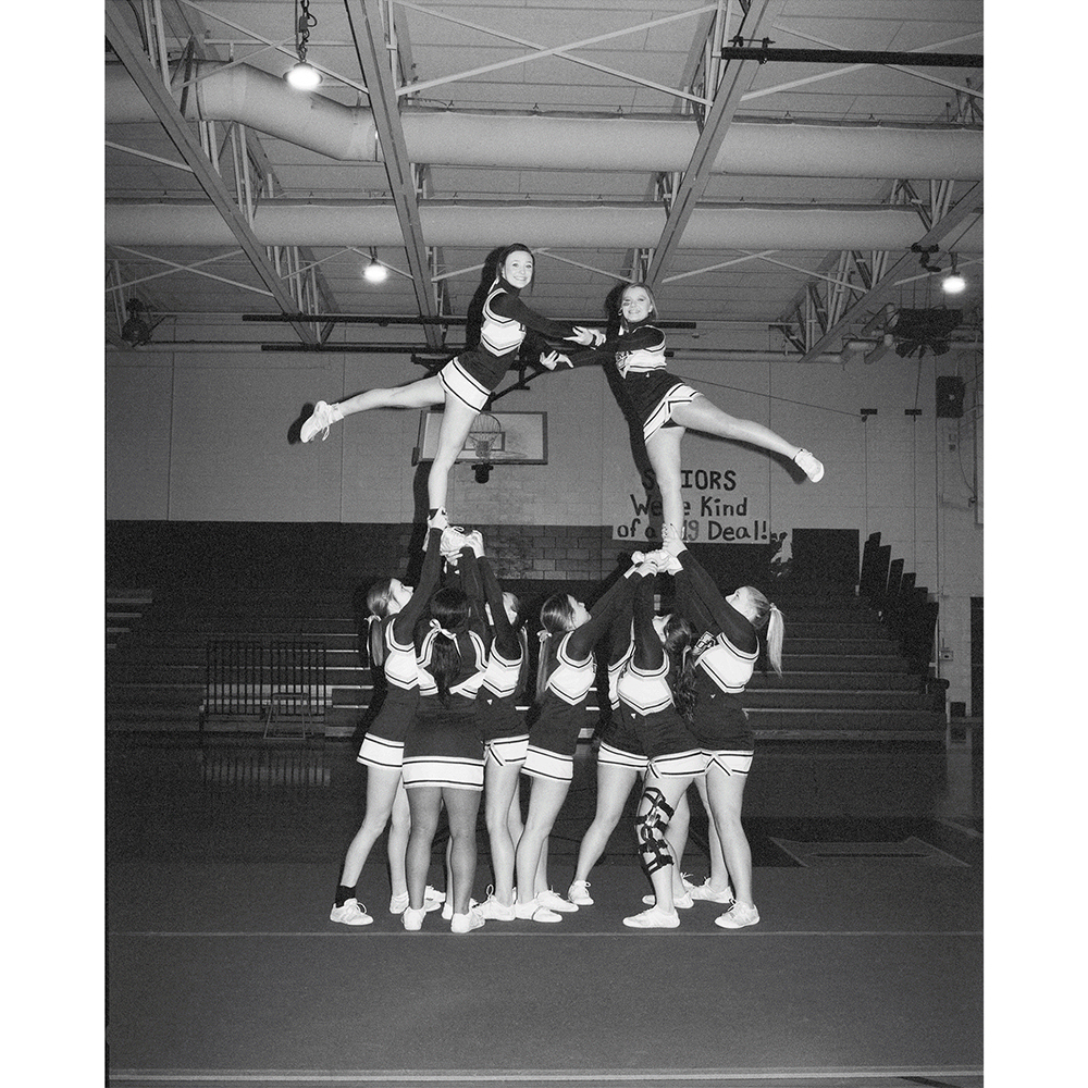 Indoor cheerleading team performing a stunt with two cheerleaders elevated by supporting teammates, classic monochrome photo.