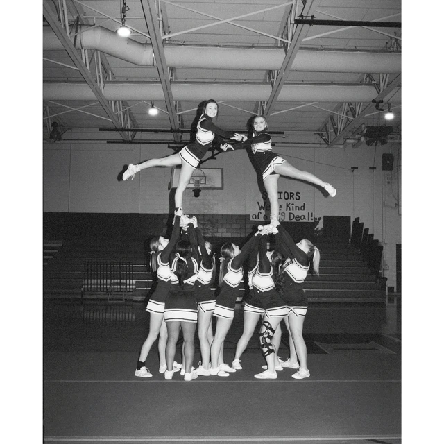 Indoor cheerleading team performing a stunt with two cheerleaders elevated by supporting teammates, classic monochrome photo.