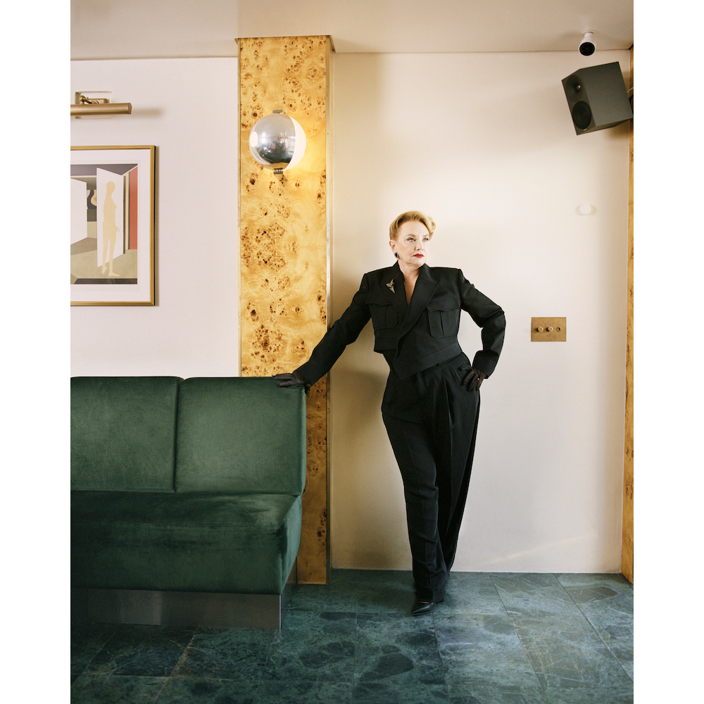 Indoor portrait of a mature woman in a black suit leaning against a wall, featuring a green couch, artwork, and disco ball lighting.