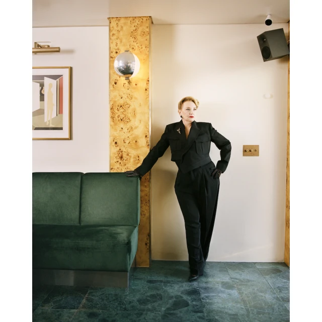 Indoor portrait of a mature woman in a black suit leaning against a wall, featuring a green couch, artwork, and disco ball lighting.
