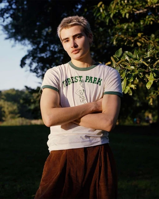 Portrait of a blonde adult male with arms crossed, wearing a graphic tee, in a verdant outdoor setting with trees and green grass.