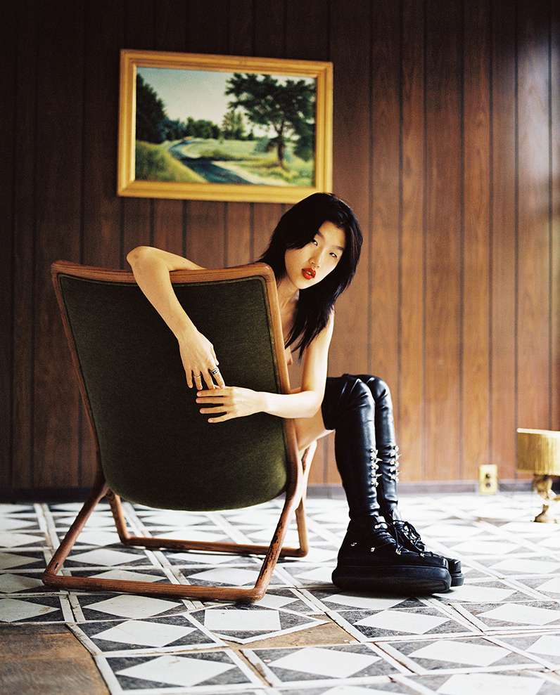 Asian woman in a green chair poses in an indoor setting with wood paneling, wearing a white top and black platform boots.