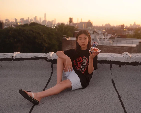 Young person sitting on a rooftop at sunset holding a small cake with candles, with a city skyline visible in the distance.