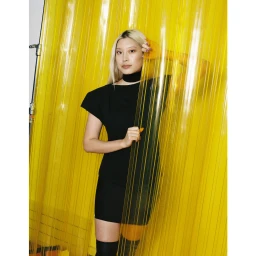 Blonde woman wearing a black dress and choker standing behind a translucent yellow plastic strip curtain.