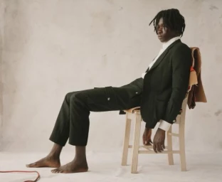 Black man with dreadlocks wearing dark green suit and white shirt sitting on a wooden chair against a pale wall.