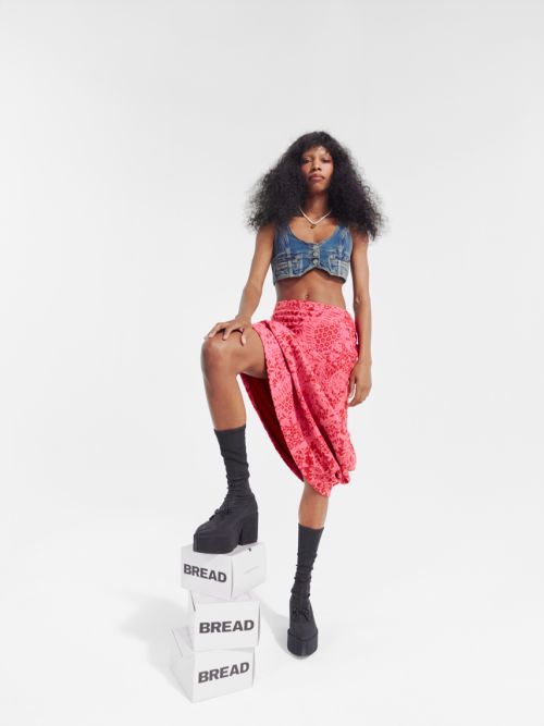 A woman in a denim top, pink skirt, platform boots and socks stands on three blocks labeled 'Bread' in a stark white studio.