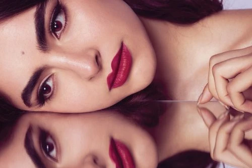 Close-up portrait of a woman with bold red lipstick and defined eyebrows reflected in a mirror surface