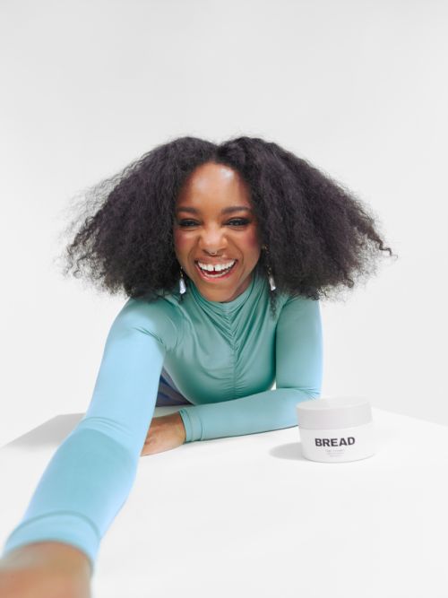 Cheerful African-American woman poses with natural hair and a turquoise shirt. She is smiling next to a jar of hair product against a white backdrop.