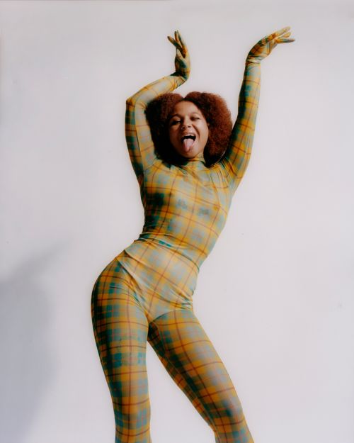 Young woman in a plaid bodysuit strikes a playful pose with hands raised and tongue out against a plain white background in a studio setting.