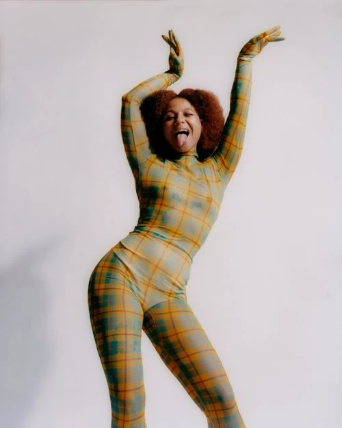 Young woman in a plaid bodysuit strikes a playful pose with hands raised and tongue out against a plain white background in a studio setting.