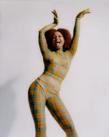 Young woman in a plaid bodysuit strikes a playful pose with hands raised and tongue out against a plain white background in a studio setting.