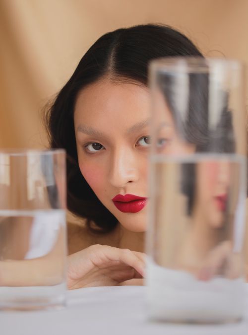 Young Asian woman with dark hair and bold red lipstick poses behind multiple glasses of water in a beauty portrait, lit by studio lighting.