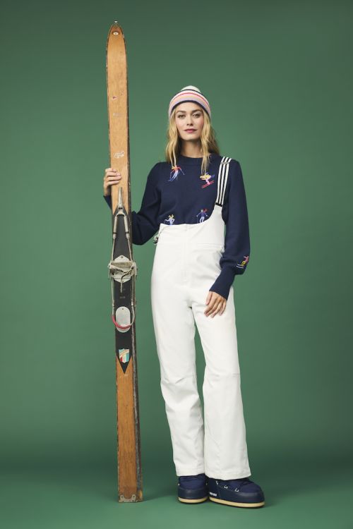 Woman poses with vintage skis in navy sweater with skier design, white overalls, and blue moon boots against a green backdrop.