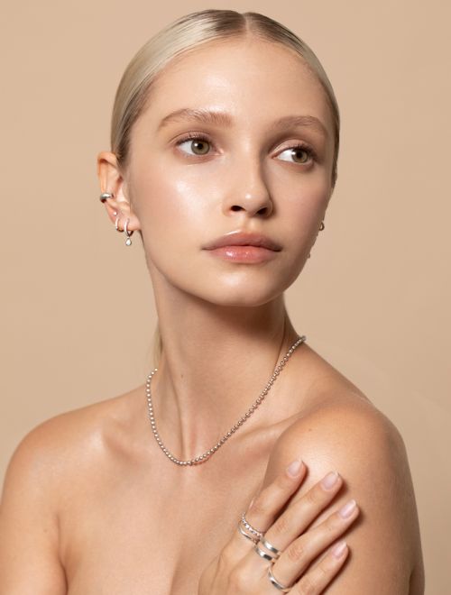 Close-up of a blonde-haired woman with bare shoulders wearing a silver necklace and rings against a beige background.