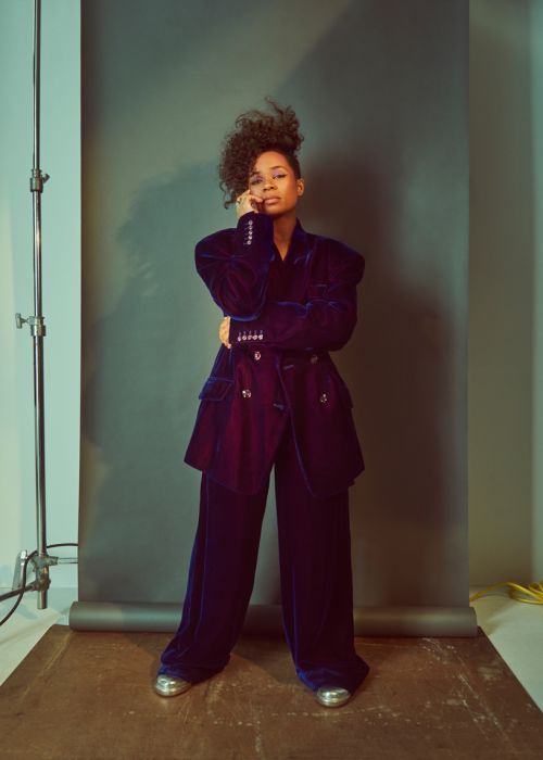 Full-length portrait of woman with curly hair in velvet blazer and pants suit with shiny silver shoes against a grey backdrop.