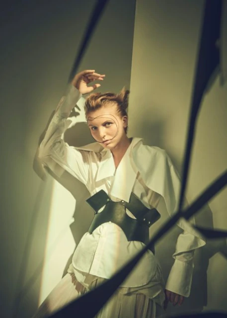 A woman with hair styled in pointed tufts, wearing a white button-down shirt and a black leather corset, poses indoors against a gray wall.