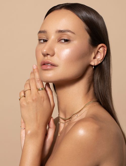 Woman with brown hair poses in a close-up studio shot wearing gold jewelry and rings against a tan background, looking at camera.