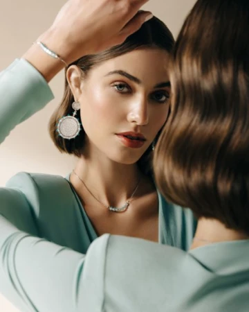 Close-up portrait of a woman wearing a mint green blazer, statement earrings, necklace, and bracelet reflected in a mirror.