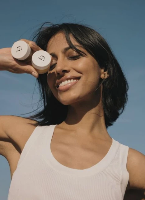 Brunette woman with beaming smile holds two small cosmetic containers against blue sky. Beauty product advertising shot with natural lighting.